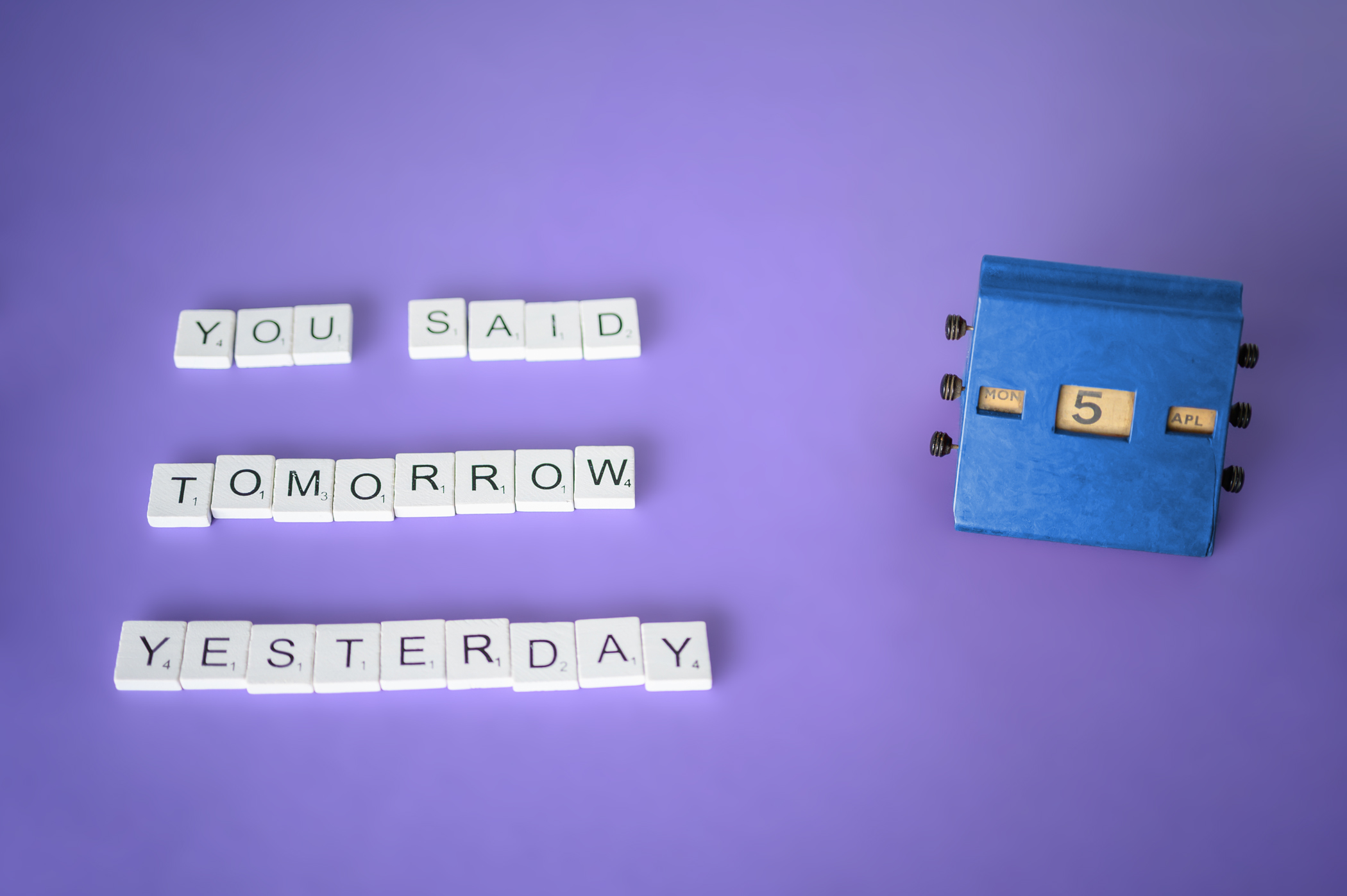 Letter tiles on a purole background spelling "you said tomorrow yesterday" next to a desk calendar