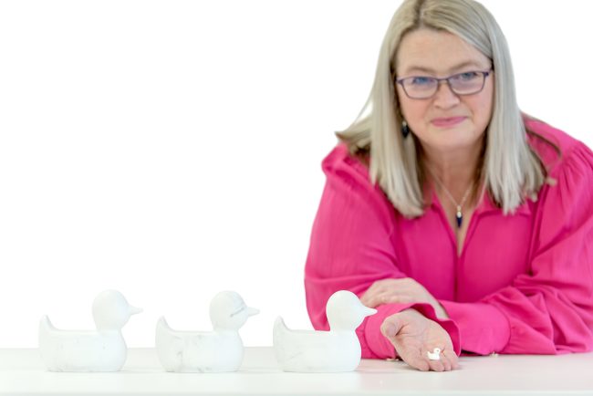 Smiling woman in a bright pink blouse holding a tiny duck figurine, with three larger ducks lined up in front, symbolising getting your ducks in a row.