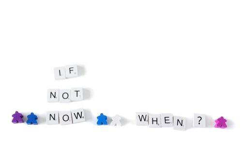 Scrabble tiles spelling out “If not now, when?” with small coloured game pieces representing people standing around the words.